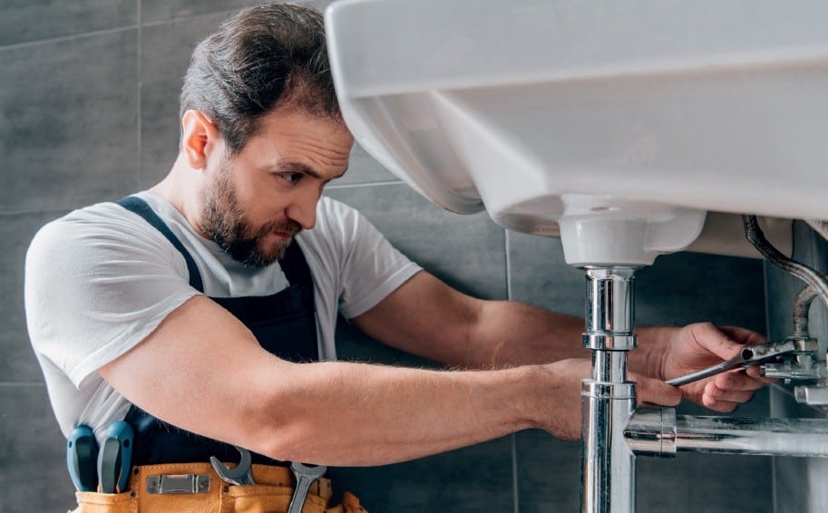 Plumber tightening bathroom sink pipes under the basin with a wrench, wearing a tool belt.